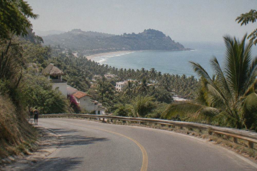 A narrow coastal road curving between green palm-covered hills with a glimpse of ocean beyond