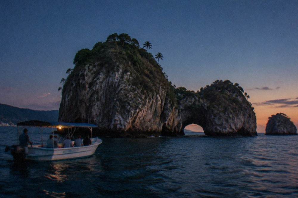 A tour boat anchored near tall rock islands in calm water as the sky turns orange at dusk