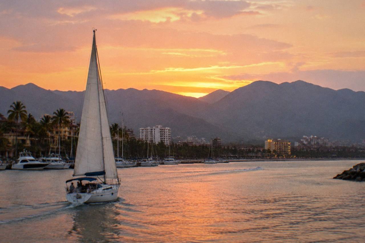 A catamaran cuts across the bay at dusk, warm light on the water and headlands beyond.